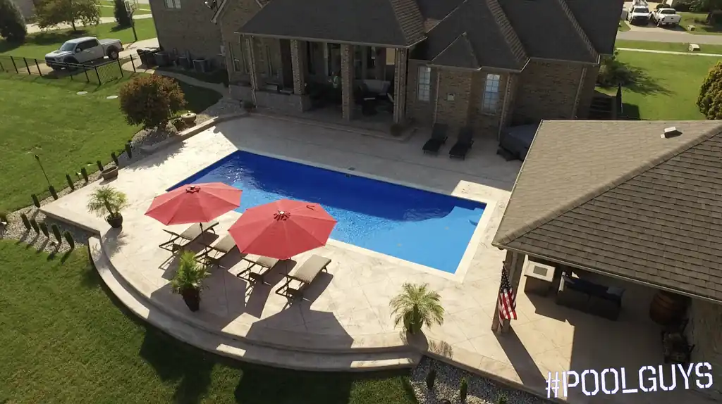 Pool with a curved patio and red umbrellas between a house and a pool house