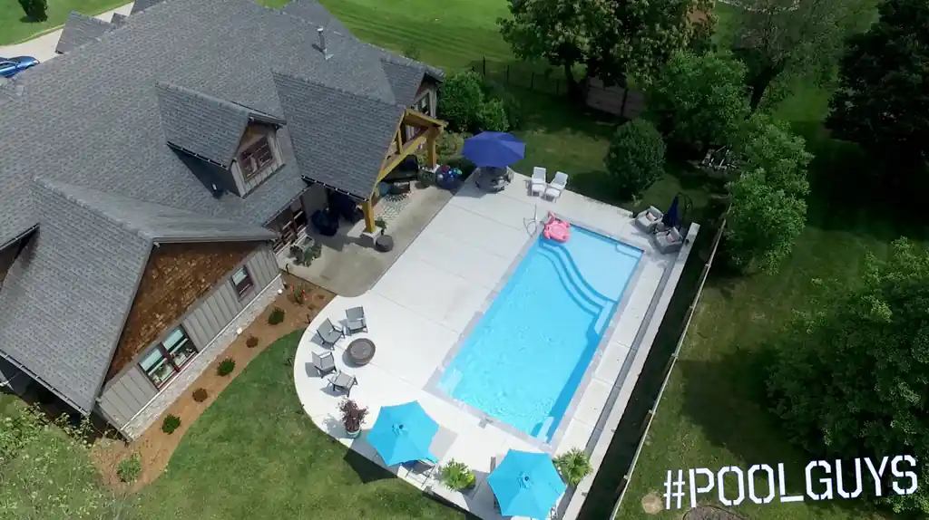Drone view of a pool with blue umbrellas and a wide concrete deck behind a shingle-sided house