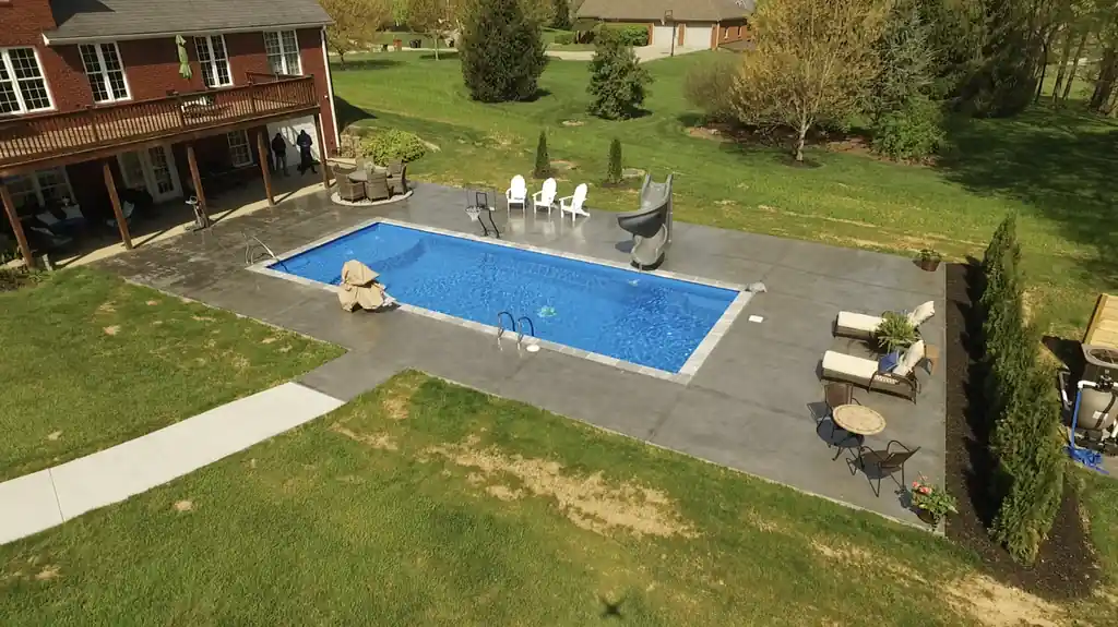 Rectangular pool with a slide on a grey concrete deck behind a brick house, open lawn beyond