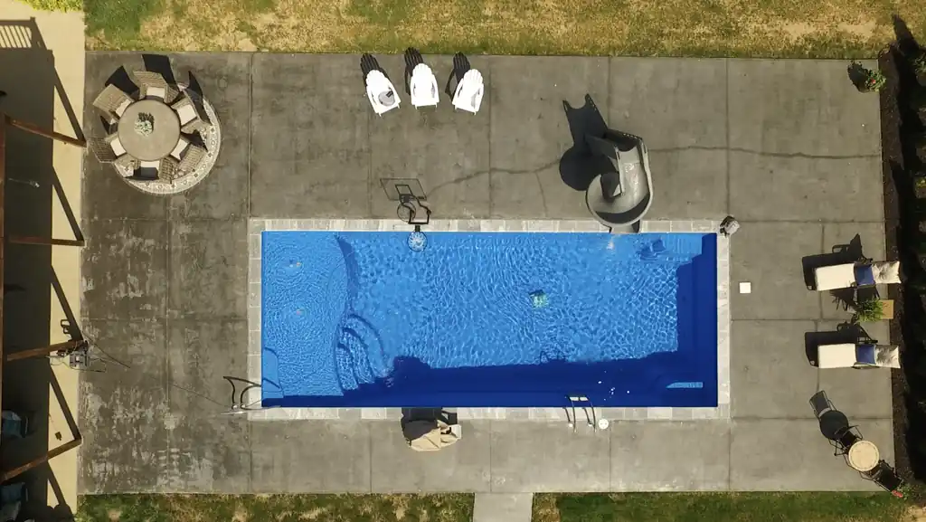 Straight-down drone view of a rectangular pool with a slide, entry steps and Adirondack chairs on a scored concrete deck