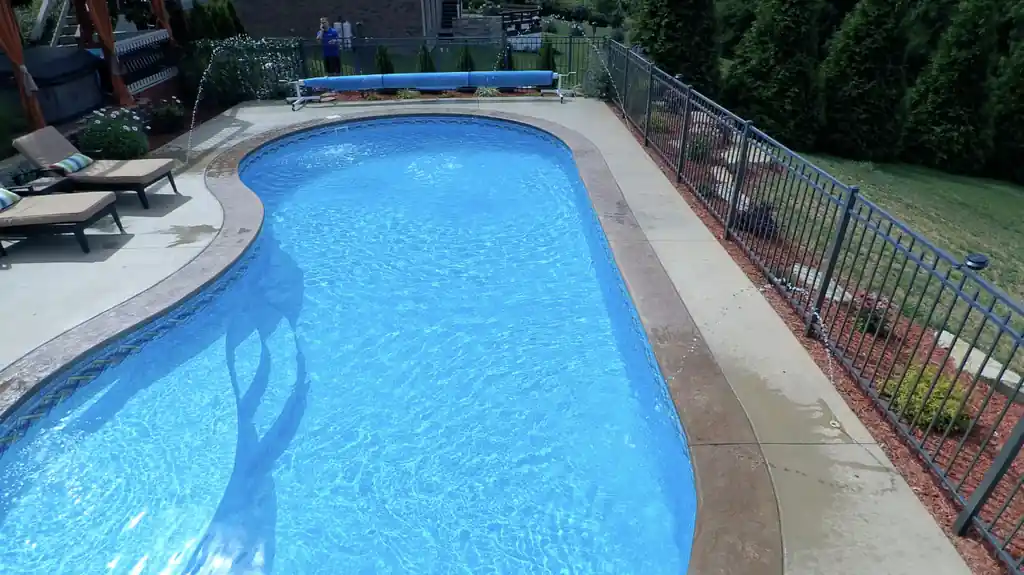 Blue pool with deck jets arcing in, running beside a black aluminium fence
